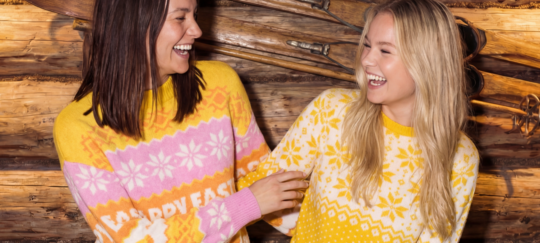 Twee lachende vrouwen in vrolijke geel-roze paastruien voor een houten wand met ski's
