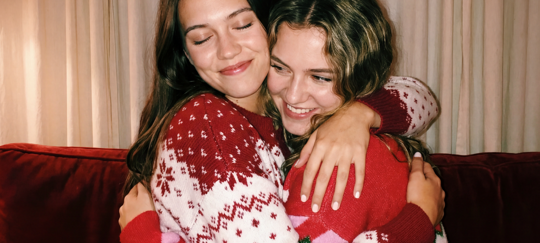 Twee lachende vrouwen knuffelen op een rode bank in gebreide kersttruien met Noors sneeuwvlokpatroon
